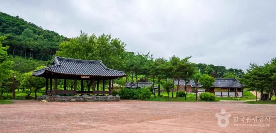 넓은 흙마당 한쪽에 기와지붕 정자가 서 있고 뒤로 나무가 보이는 모습