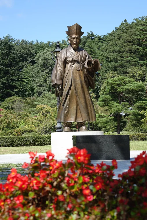 붉은 꽃밭 뒤 받침대 위에 선 율곡 이이 동상