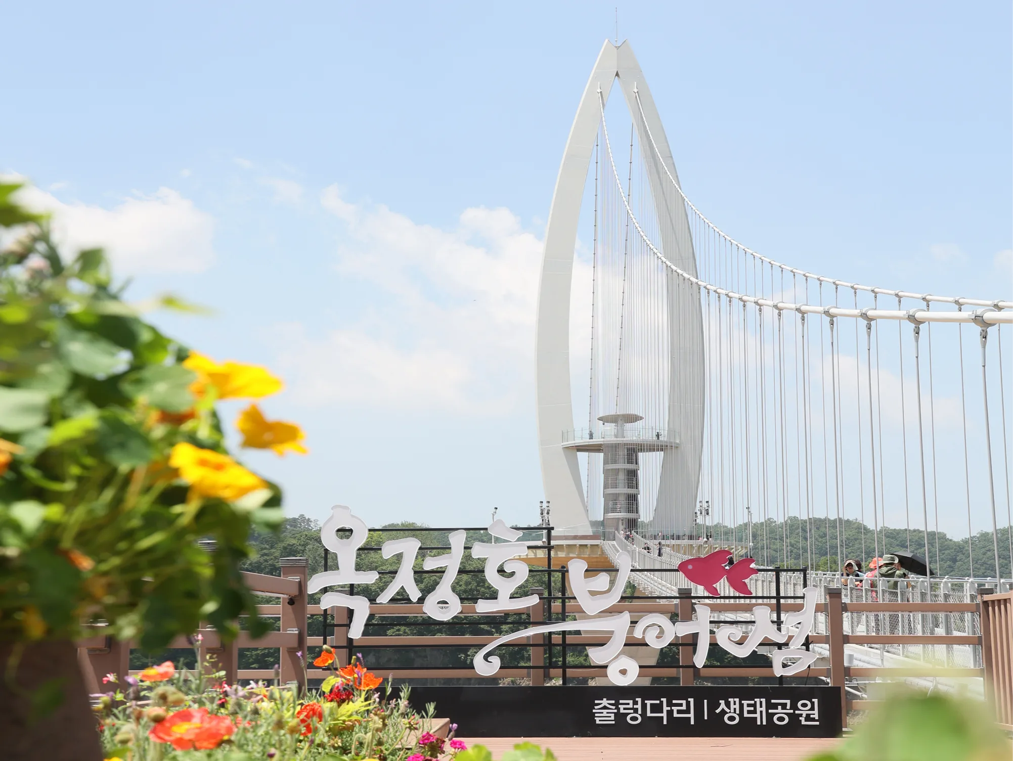 옥정호 붕어섬 출렁다리 입구의 조형 표지판과 꽃, 다리를 건너는 방문객들