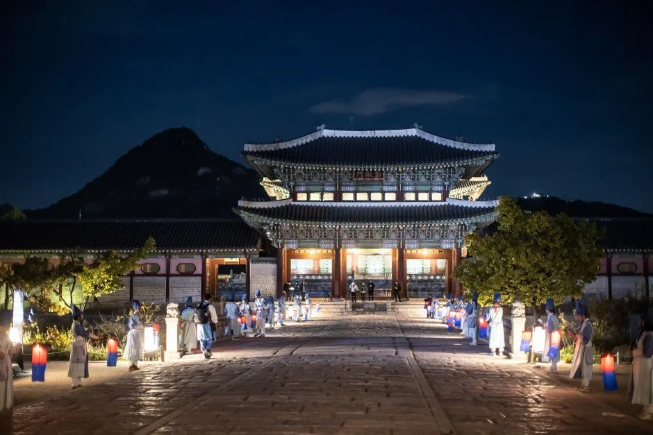 밤에 등불이 늘어선 돌길 끝에 조명을 받은 2층 지붕 전각이 서 있고 뒤로 산 능선이 보이는 궁궐 야경