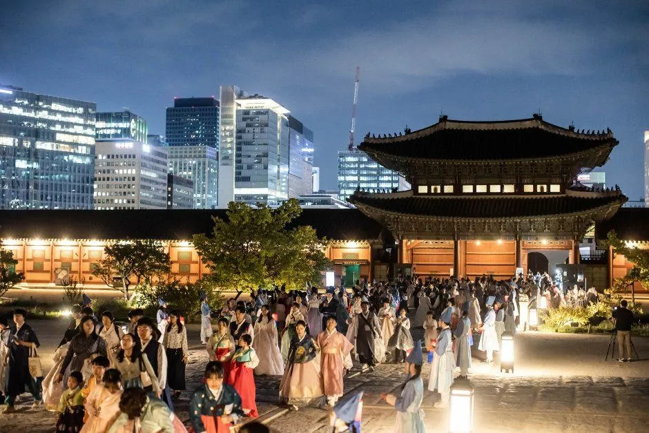 야간 조명이 켜진 궁궐 전각 앞마당에 한복 차림을 포함한 많은 관람객이 모여 있고 뒤로 고층 건물이 보이는 풍경