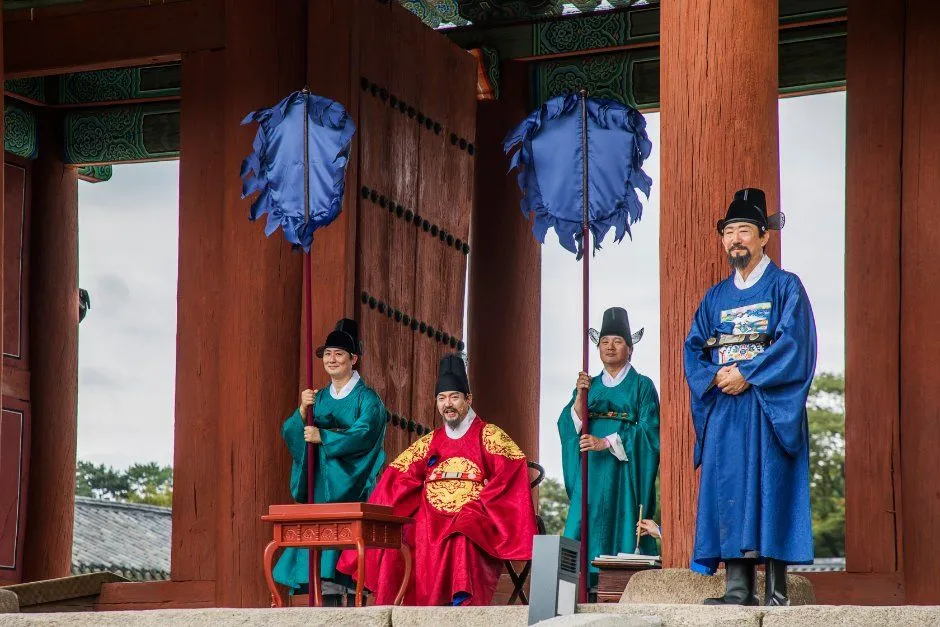 붉은 기둥이 서 있는 문 앞에서 붉은 곤룡포 차림 인물과 관복 차림 인물들이 앉거나 서 있는 왕실 재현 장면