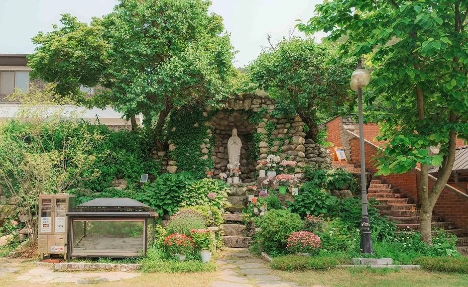 돌로 쌓은 감실과 성모상, 꽃과 나무가 있는 성당 경내 정원