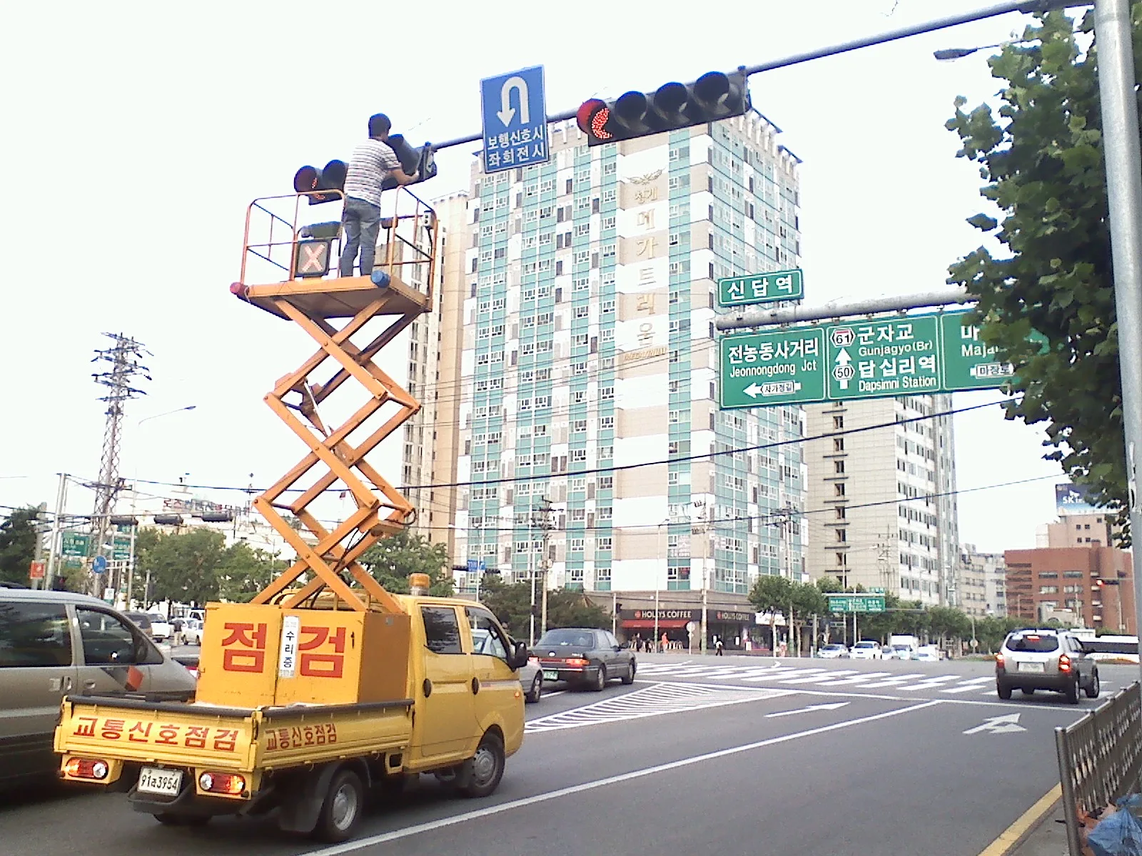 고소작업차에 오른 작업자가 교차로 신호등을 점검·정비하는 모습, 신호등에는 적색불이 켜져 있고 도로에는 차량들이 오가고 있다