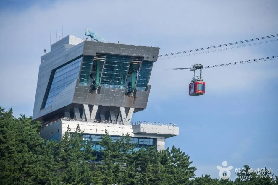 바다 옆 산자락에 세워진 케이블카 역사 건물과 붉은 캐빈이 지나가는 모습