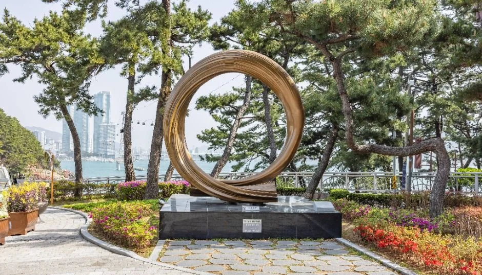 원형 조형물과 꽃이 핀 산책로, 멀리 도심 건물이 보이는 공원길