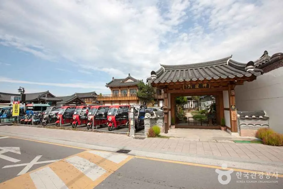 한옥 기와지붕 건물과 가게들이 이어진 낮의 거리 한쪽에 붉은 소형 전동차들이 주차되어 있는 모습