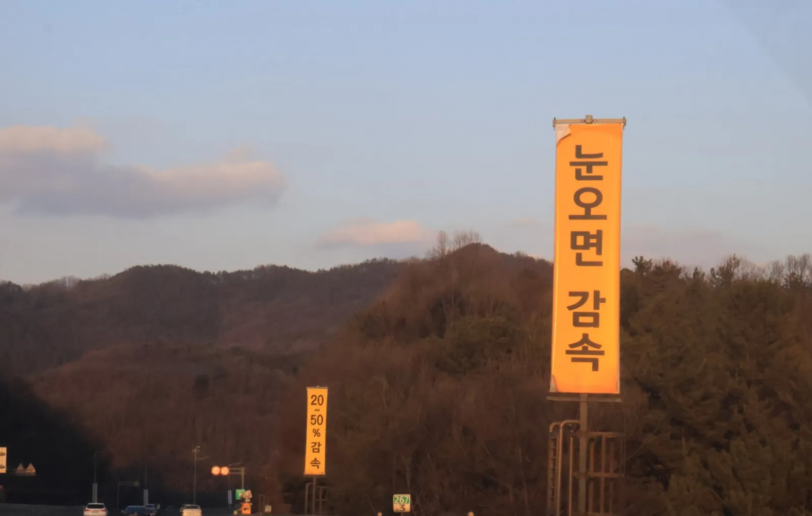 노을빛이 남은 하늘 아래 산을 배경으로 한 도로 위 "눈오면 감속" 현수막이 걸려 있는 모습