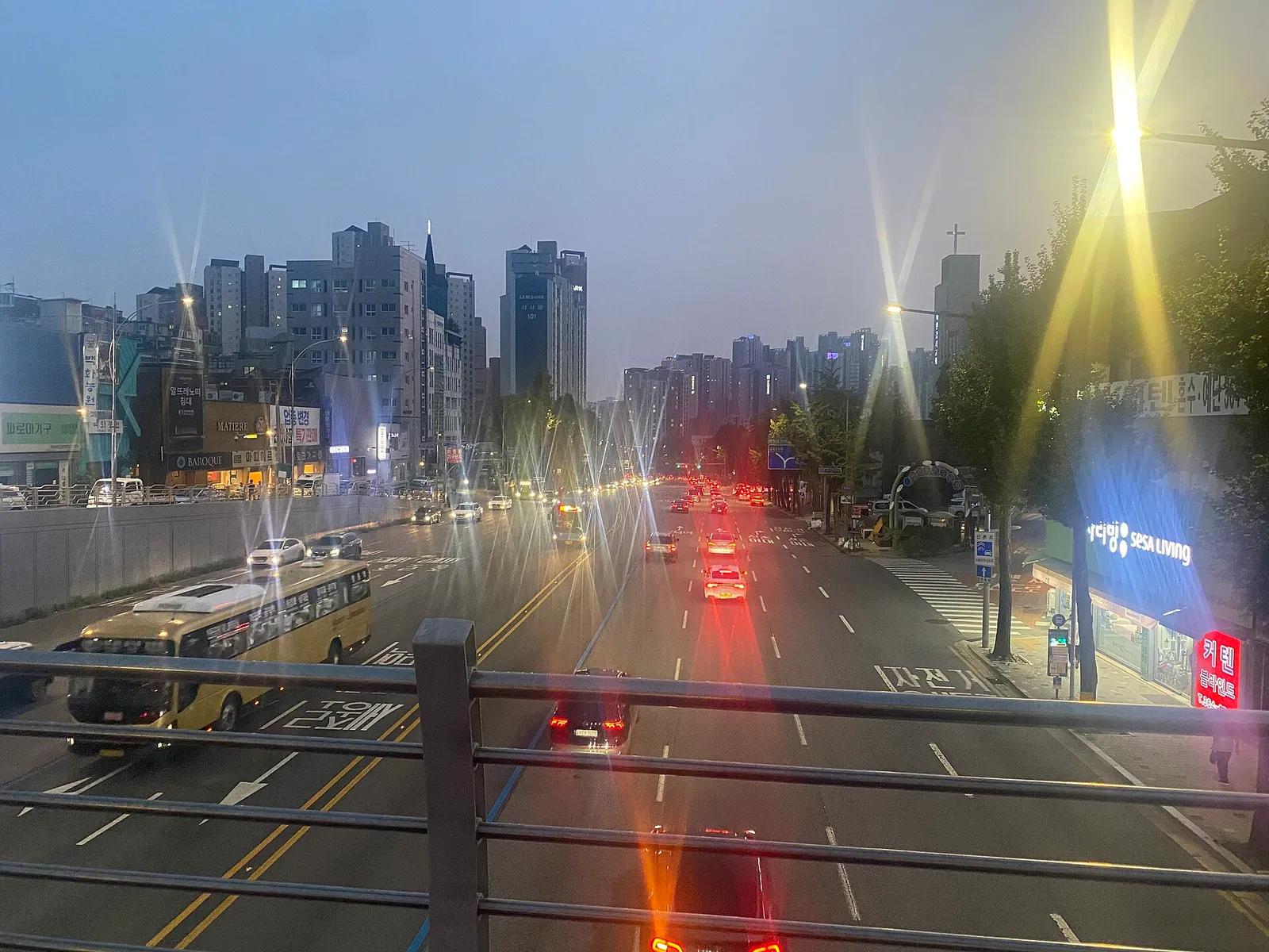 서울 도심 도로에서 해 질 무렵 버스와 승용차들이 전조등과 브레이크등을 켠 채 달리고, 가로등 빛이 번져 보이는 푸른 저녁 하늘