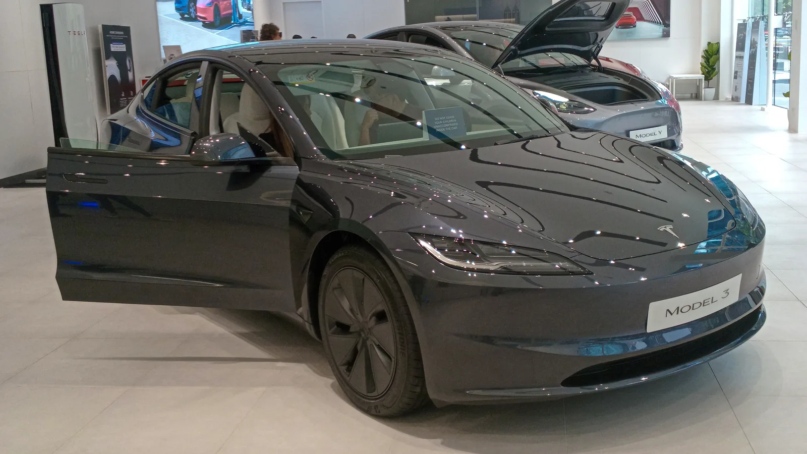 회색 테슬라 모델3가 전시장에 놓여 있고 운전석 문이 열려 있다, 앞범퍼에 MODEL 3 레터링이 보인다