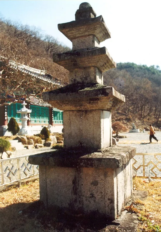 잎이 진 나무와 산을 배경으로 이끼 낀 3층 석탑이 서 있는 모습