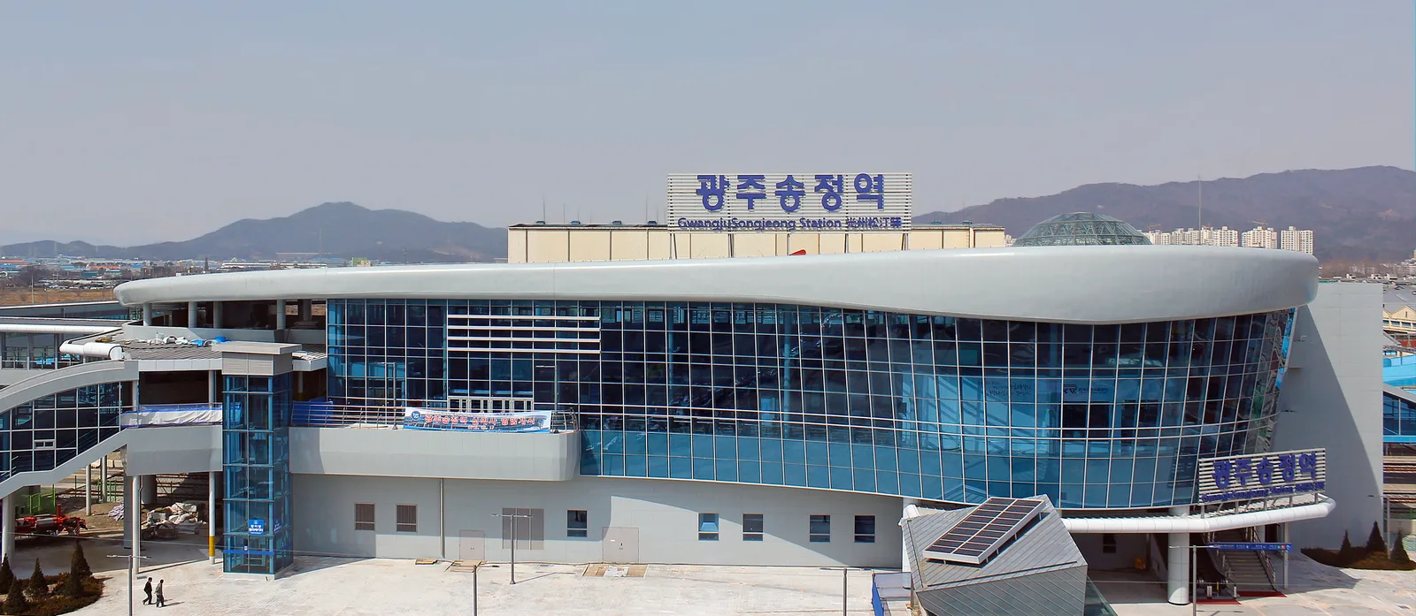 곡선형 유리 외벽을 가진 현대적인 기차역 건물, 지붕 위에 파란 글씨로 역명이 적힌 간판이 서 있다