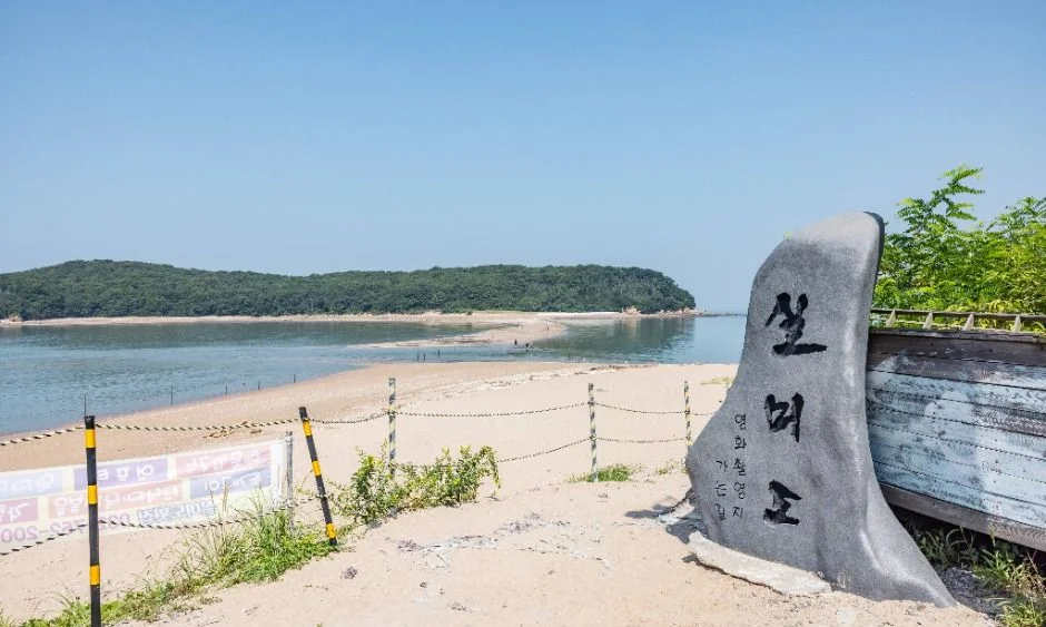 실미도라고 새긴 표지석과 그 뒤로 숲이 우거진 섬이 보이는 모래사장